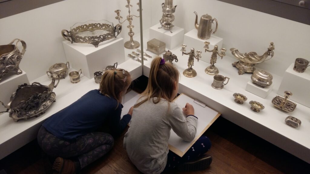Dwie, kilkuletnie dziewczynki przyglądają się srebrnym przedmiotom w szklanej gablocie. Każda ma białą kartkę i ołówek. Przygotowują się do rysowania.