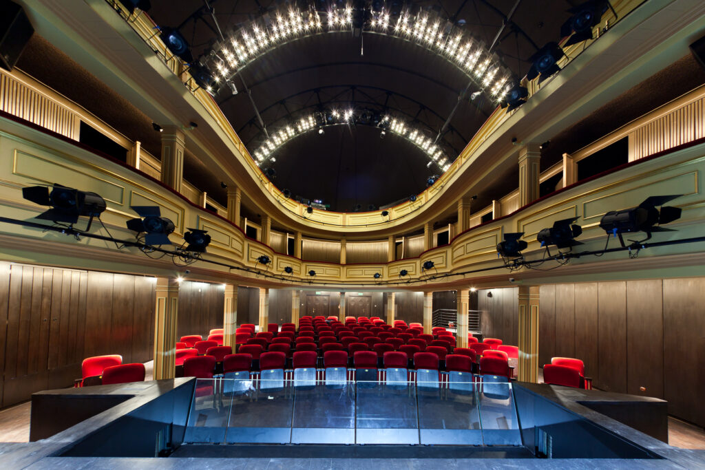 Widok na salę widowiskową Teatru Starego z poziom sceny; na pierwszym planie puste rzędy krzeseł widowni, w tle loże i balkony.