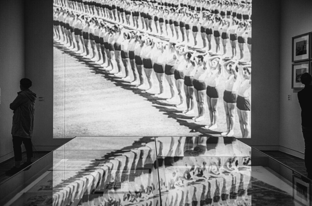 Czarno-białe zdjęcie: widok wystawy; na ścianie projekcja z rzędami gimnastyczek; ten sam obraz odbija się w szklanej gablocie.