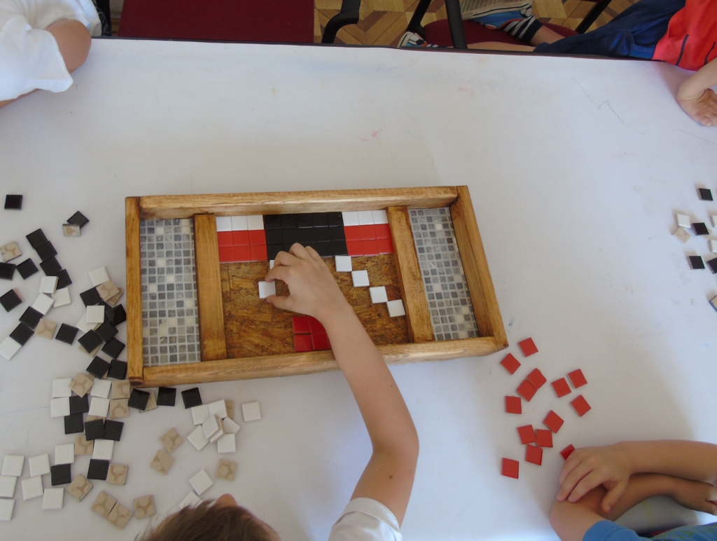 Zdjęcie. Drewniany pojemnik do układania mozaiki z kolorowych kwadratów. Biały stół, na mim rozrzucone fragmenty mozaiki. Ręce dzieci układających mozaikę.